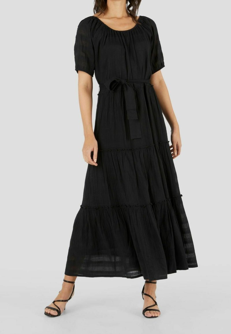Femme portant une robe longue noire à étages, manches courtes, col rond et ceinture à la taille, associée à des sandales noires à brides.