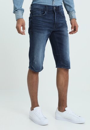 Bermuda en denim de teinte foncée avec des effets légèrement usés, arrivant à mi-cuisse, assortis à des baskets blanches.