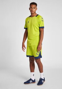 Neongrön sportströja med blå detaljer, korta ärmar och matchande shorts med en logotyp och chevronmönster; bärs med blå sneakers.