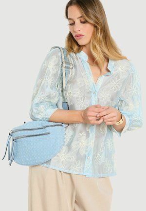 Frau trägt hellblaue, floral bestickte Bluse und beige Hose, hält eine strukturierte hellblaue Handtasche mit doppeltem Reißverschluss.