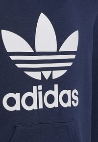 Felpa blu navy con un grande logo Adidas bianco che presenta un design a trifoglio e tre strisce orizzontali sopra il testo "adidas." Texture morbida.