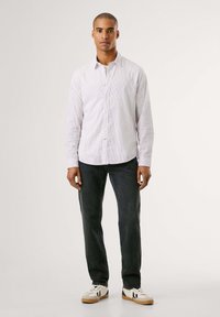 Camisa de hombre de manga larga en rosa claro con rayas verticales blancas, abotonada, llevada con jeans de mezclilla oscura y zapatillas beige.