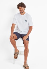 White cotton t-shirt with short sleeves and blue graphic design, paired with navy shorts and brown sandals. Model seated on a ladder.
