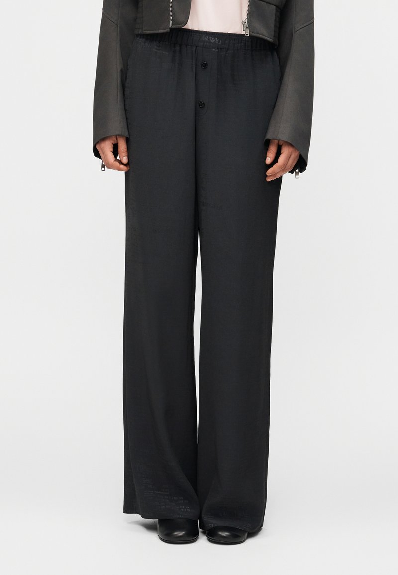 Άτομο που φοράει φαρδιά μαύρα παντ ελόνια με κουμπιά μπροστά, μαύρα παπούτσια και σκούρο cropped μπουφάν πάνω από ανοιχτόχρωμο τοπ, στέκεται μπροστά σε απλό φόντο.