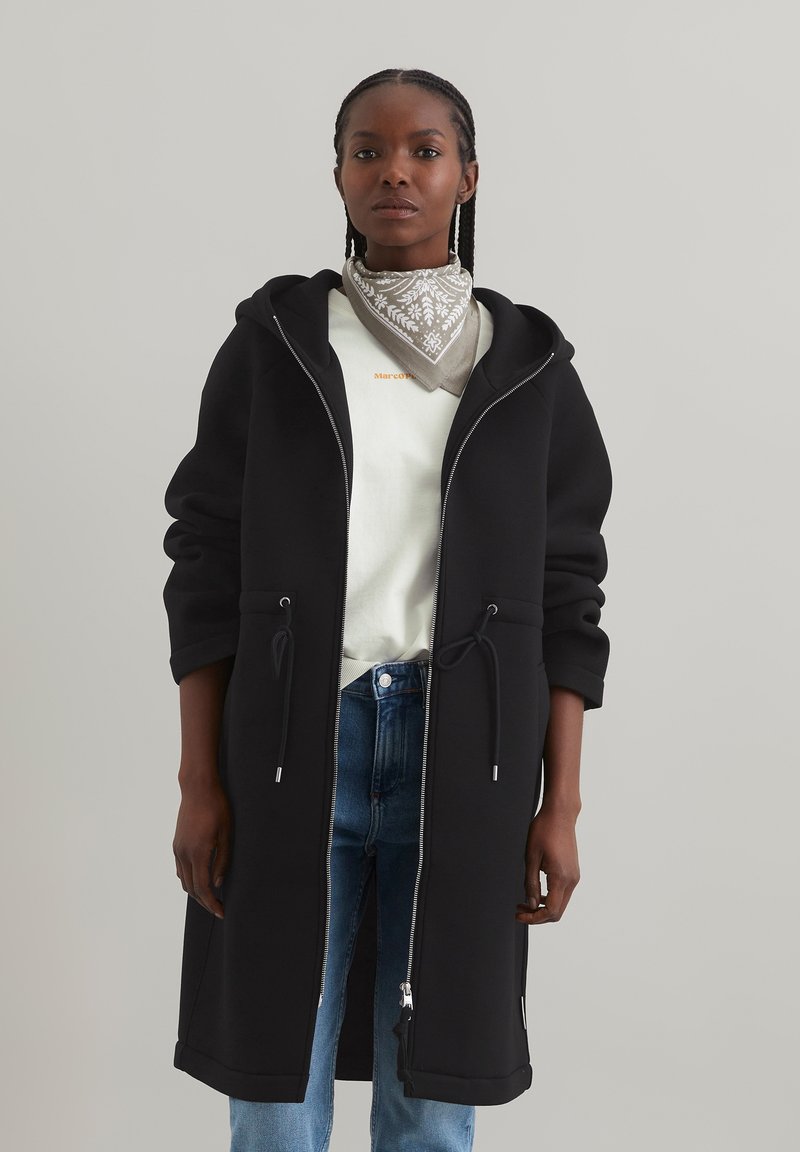 Manteau long noir à capuche en tissu lisse, doté d'une fermeture éclair sur le devant, de cordons de serrage à la taille et de manches relaxées. Porté par-dessus un haut léger.