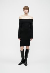 Kvinne som har på seg en svart av skulderen, knelång kjole og svarte knelengde støvler, stående mot en ensfarget bakgrunn.