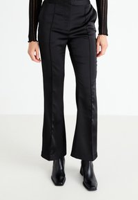 Schwarze satinierte ausgestellte Hose mit glatter Oberfläche, ausgestattet mit maßgeschneiderten Nähten und einem hochgeschnittenen Design, kombiniert mit spitzen schwarzen Ankle-Boots.