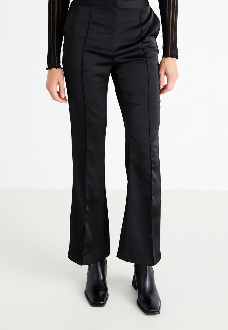 Schwarze satinierte ausgestellte Hose mit glatter Oberfläche, ausgestattet mit maßgeschneiderten Nähten und einem hochgeschnittenen Design, kombiniert mit spitzen schwarzen Ankle-Boots.