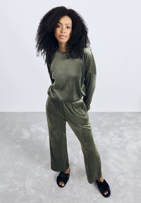 Olijfgroen fluwelen loungekleding set met losvallende lange mouwen en broeken. Zwarte fuzzy slippers. Zachte textuur tegen een lichte achtergrond.