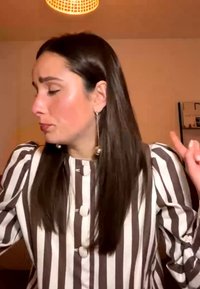 Woman with long dark hair and striped shirt gesturing with both hands in a warmly lit room with a hanging lamp above.