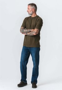 Oliven grøn T-shirt med korte ærmer, parret med blå denim jeans. Bemærkelsesværdige tatoveringer synlige på armene; brune sko fuldender looket.