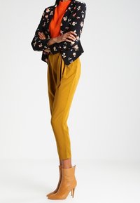 Blommigt svart kavaj, orange tröja och senapsgula byxor; beigefärgade ankelboots med avsmalnande klack; åtsittande siluett, slät tygkvalitet.