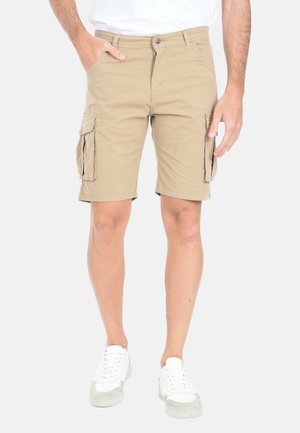 Uomo che indossa pantaloncini cargo beige con tasche a patta, scarpe da ginnastica bianche e una t-shirt bianca, in piedi davanti a uno sfondo semplice.