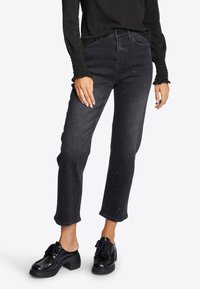 Jeans in denim nero con taglio dritto, impreziositi da piccoli chiodi in argento, indossati con scarpe nere con tacco spesso.