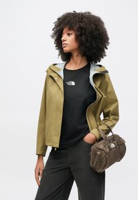 Αδιάβροχο μπουφάν σε ελαιί πράσινο με γκρι επένδυσ�η, συνδυασμένο με μαύρο t-shirt με λογότυπο, σκούρο παντελόνι και καφέ τσάντα με γούνινες λεπτομέρειες.