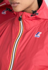 Chaqueta impermeable roja con un cuello alto, que presenta una cremallera negra y acentos de rayas coloridas. Parche del logo en la parte frontal izquierda.