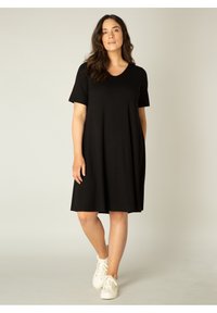 Zwart korte mouwen jurk met een V-hals, gemaakt van zachte stof, met een ontspannen pasvorm die boven de knie valt. Gemaakt met witte sneakers.