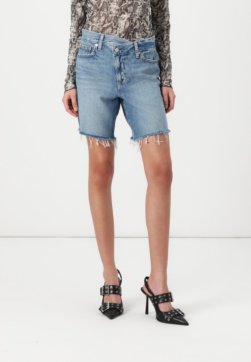 Short en jean bleu clair avec ourlets bruts, porté avec des chaussures à talons noires à brides bouclées, associé à un top à manches longues imprimé animal.