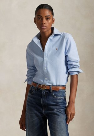 Kobieta w jasnoniebieskiej koszuli zapinanej na guziki z podwiniętymi rękawami, wpuszczona w niebieskie spodnie dżinsowe z wysokim stanem i brązowym skórzanym paskiem.