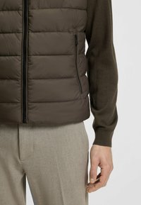 Braune gesteppte Weste mit horizontalem Steppmuster, Frontreißverschluss und seitlicher Tasche; getragen über einem dunkelbraunen Pullover und hellgrauen Hose.
