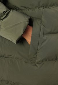 Green puffer coat material with a smooth, shiny texture. Features a large side pocket, elastic cuffs, and a flexible, padded design.