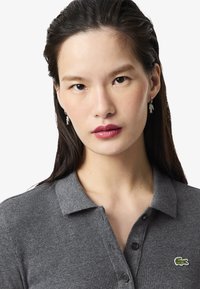 Polo gris côtelé avec un col, une patte de boutonnage et un logo Lacoste vert. Le modèle porte des boucles d'oreilles en argent en forme de poisson.
