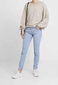 Suéter de punto beige con mangas abullonadas, jeans azul claros con puños doblados y zapatillas blancas. El conjunto es informal y ajustado.
