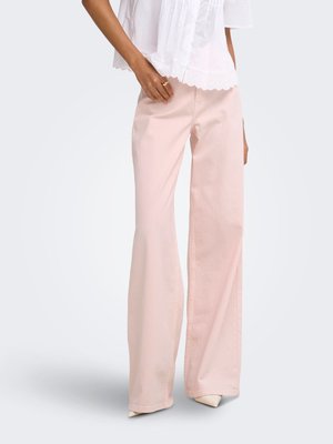 Femme portant un pantalon large rose clair associé à un chemisier blanc et des talons blancs à bouts pointus, debout devant un fond uni.