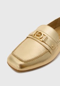 Gold loafers with a textured metallic surface, featuring a gold strap and round logo accent, square toe design, and minimal stitching.