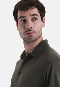 Olive zelené polo tričko s texturovaným povrchem, třemi knoflíky a límečkem. Jemný oranžový akcent viditelný na vnitřní straně límečku.