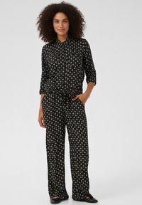 Svart jumpsuit med blommönster, knappfront, 3/4-ärm och dragsko i midjan. Har löst sittande byxor och platta skor.