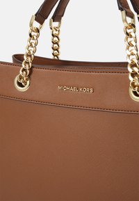 Tansyntetisk läderhandväska med en slät yta, guldkedjeaccent och en logo, med en strukturerad form och topphandtag.