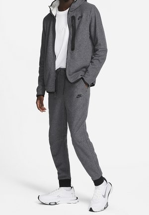 Mann trägt grauen Nike-Hoodie und Jogginghose mit weißen Nike-Sneakers, steht mit einer Hand in der Tasche vor weißem Hintergrund.