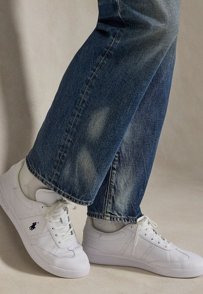 Persoon draagt witte vetersneakers met een zwart logo en blauwe spijkerbroek op een beige vloer.