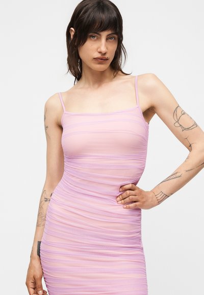 Γυναίκα με σκούρα μαλλιά και τατουάζ στα χέρια, φοράει στενό, ριγέ λιλά φόρεμα με λεπτές τιράντες, χέρι στη μέση, ουδέτερο φόντο.