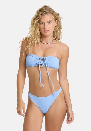 Femme blonde aux cheveux bouclés portant un bikini bandeau bleu clair et un collier de perles coloré, debout devant un fond uni.