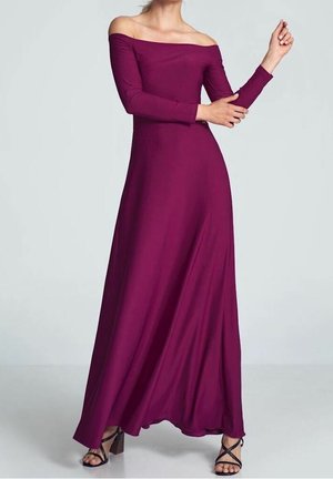 Frau in einem bodenlangen, schulterfreien magentafarbenen Kleid mit langen Ärmeln und schwarzen, hochhackigen Sandalen mit Riemchen, stehend vor einem grauen Hintergrund.