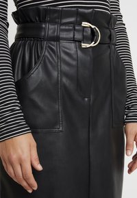 Svart fusk läder kjol med en bred midjeband och ett framsidan spänne detaljer, två sidofickor och en slät yta. Randig toppärm synlig.