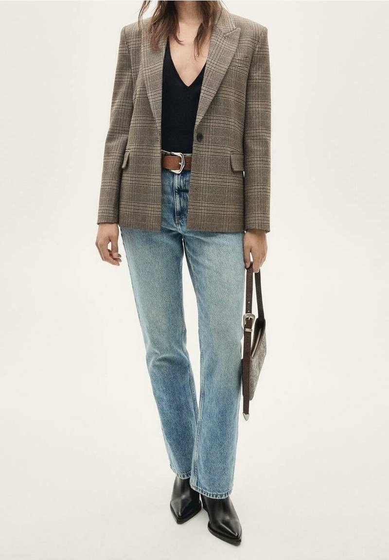 Femme portant un blazer marron à carreaux, un top noir, un jean bleu clair, des bottes noires pointues, tenant un sac à main sombre à ses côtés.