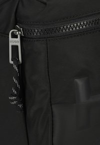 Zaino in tessuto nero con un'esterno strutturato, dotato di una cerniera metallica con tiralampo e una patch in materiale liscio a contrasto.