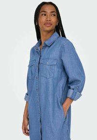 Denim haljina u svijetloplavoj boji, s dugmadi sprijeda, dva džepa na prsima i podvrnutim rukavima. Mekana tekstura, opušteni kroj.