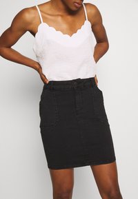 Weißes, mit Wellenrand versehenes Tanktop, kombiniert mit einem hochgeschnittenen schwarzen Jeansrock, der über Fronttaschen und eine taillierte Form verfügt.