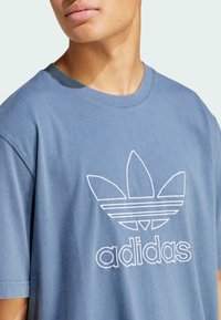 Niebieska bawełniana koszulka z białym haftowanym logo Adidasa z motywem liścia i poziomymi paskami, okrągły dekolt, krótkie rękawy.
