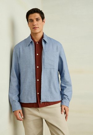 Young man wearing a light blue zip-up jacket over a brown button-up shirt and beige pants, standing against a plain light wall.