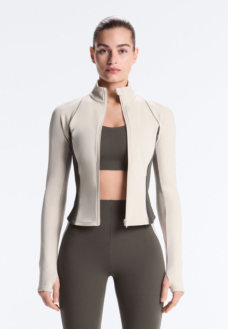 Femeie purtând o jachetă bej strâmtă cu fermoar peste un sutien sportiv închis la culoare și colanți cu talie înaltă, stând pe un fundal alb.