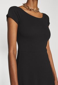 Svart, räfflad klänning med korta ärmar och rund halsringning. Accentuerad med ett kraftigt guldhalsband. Slät textur.