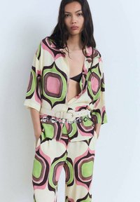 Gemusterter Kleidungssatz mit einem kurzärmeligen Hemd und einer Hose. Die Farben umfassen Creme, Rosa, Grün und Braun mit geometrischen Formen. Gürtel mit kreisförmigen Akzenten.