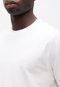 Κοντινή λήψη ενός ατόμου που φοράει ένα απλό λευκό μπλουζάκι με στρογγυλή λαιμόκοψη και ένα μικρό, διακριτικό λογότυπο "ck" κεντημένο στο στήθος.