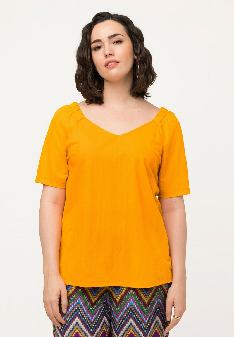 Top de manga corta con escote en V en tejido amarillo brillante de textura suave y detalle fruncido en el escote, combinado con pantalones estampados.