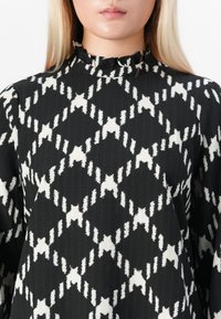 Donna che indossa una blusa nera con motivo geometrico a diamante bianco e un piccolo collo alto arricciato, vista dalle spalle al mento.